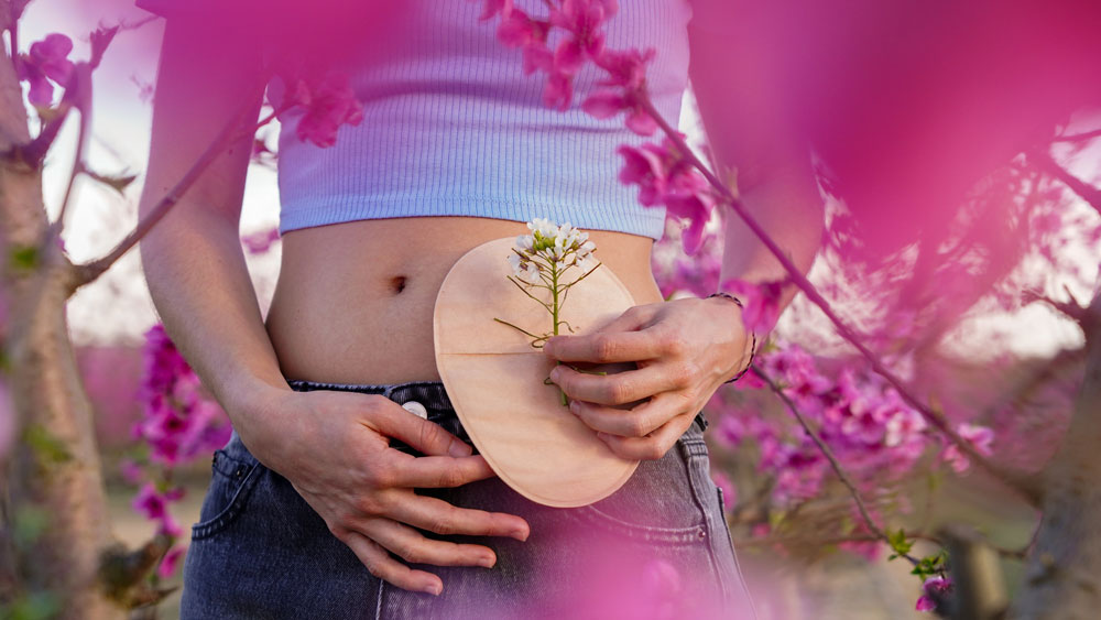 une femme tient d'une main un sac de stomie et une fleur et de l'autre main, elle entoure son bacuh inférieur. A l'arrière-plan, on voit des fleurs et une prairie.